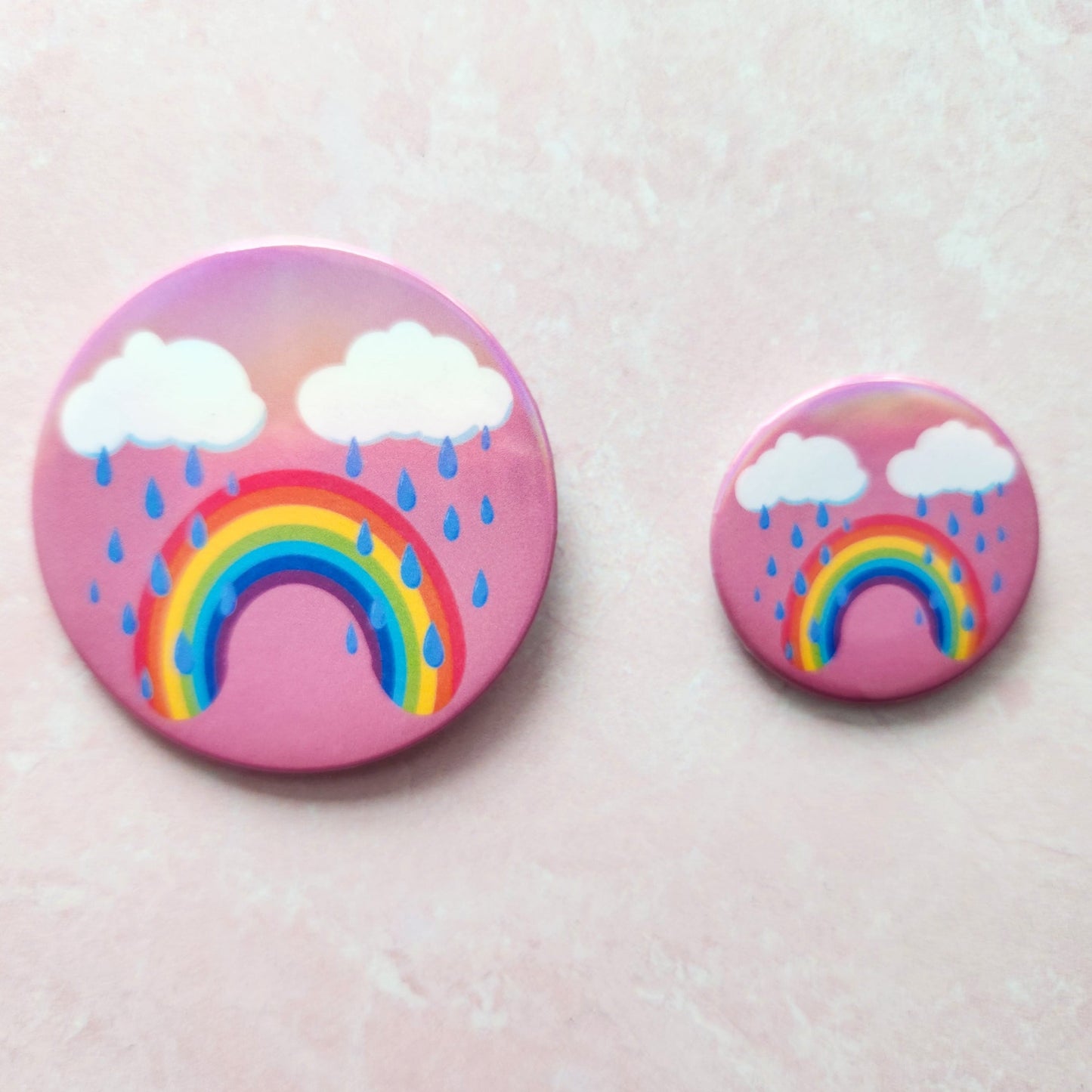 Bittersweet rainbow holographic button pin with a pink background, featuring a crying rainbow under two clouds with raindrops, one larger next to a smaller