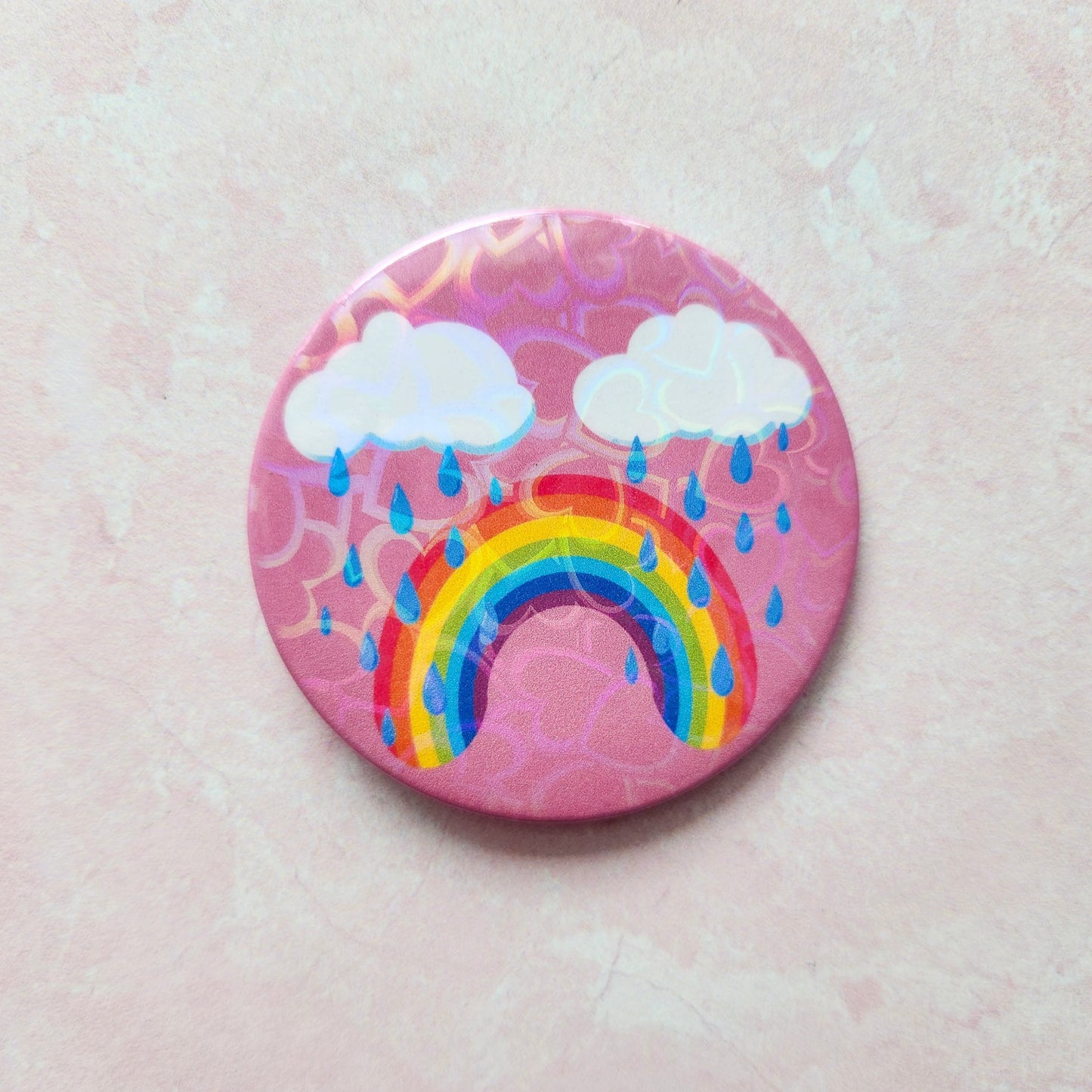 Bittersweet rainbow holographic button pin with a pink background, featuring a crying rainbow under two clouds with raindrops
