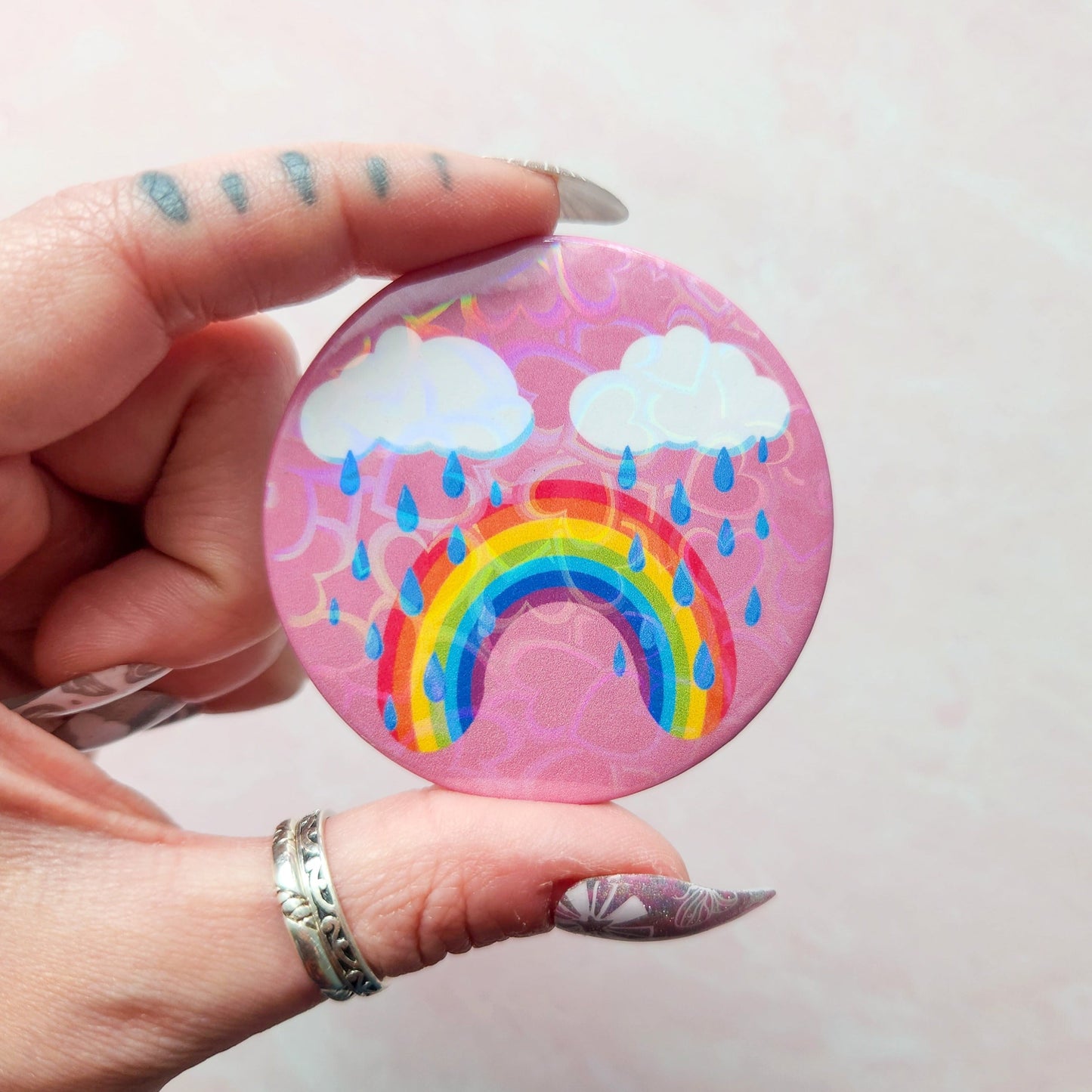 Bittersweet rainbow holographic button pin with a pink background, featuring a crying rainbow under two clouds with raindrops, held between fingers with decorative nail art.