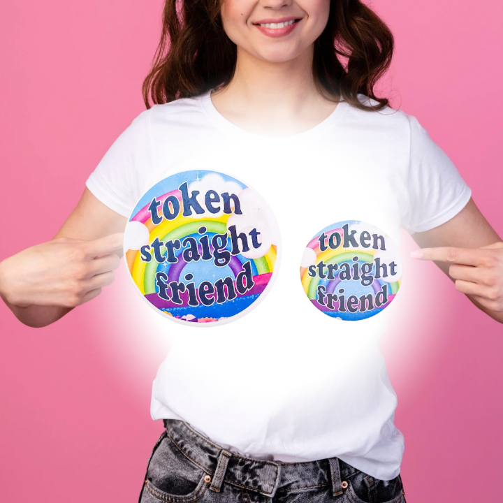 Person wearing a pink shirt with a close-up view of two sizes of colorful 'Token Straight Friend' holographic pin featuring a bold rainbow design and playful text, perfect for pride events and allyship.