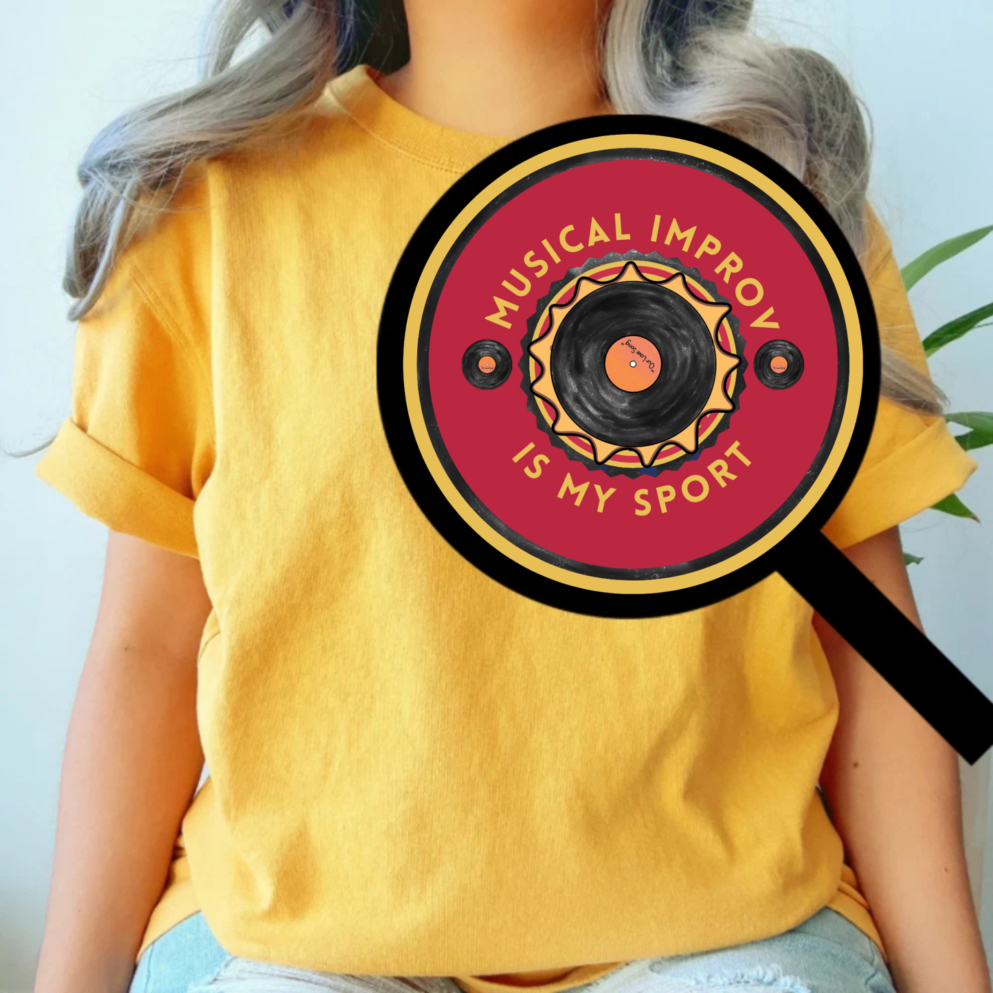 Person in yellow shirt wearing a musical improv is my sport pin back button, magnified so we can see it.