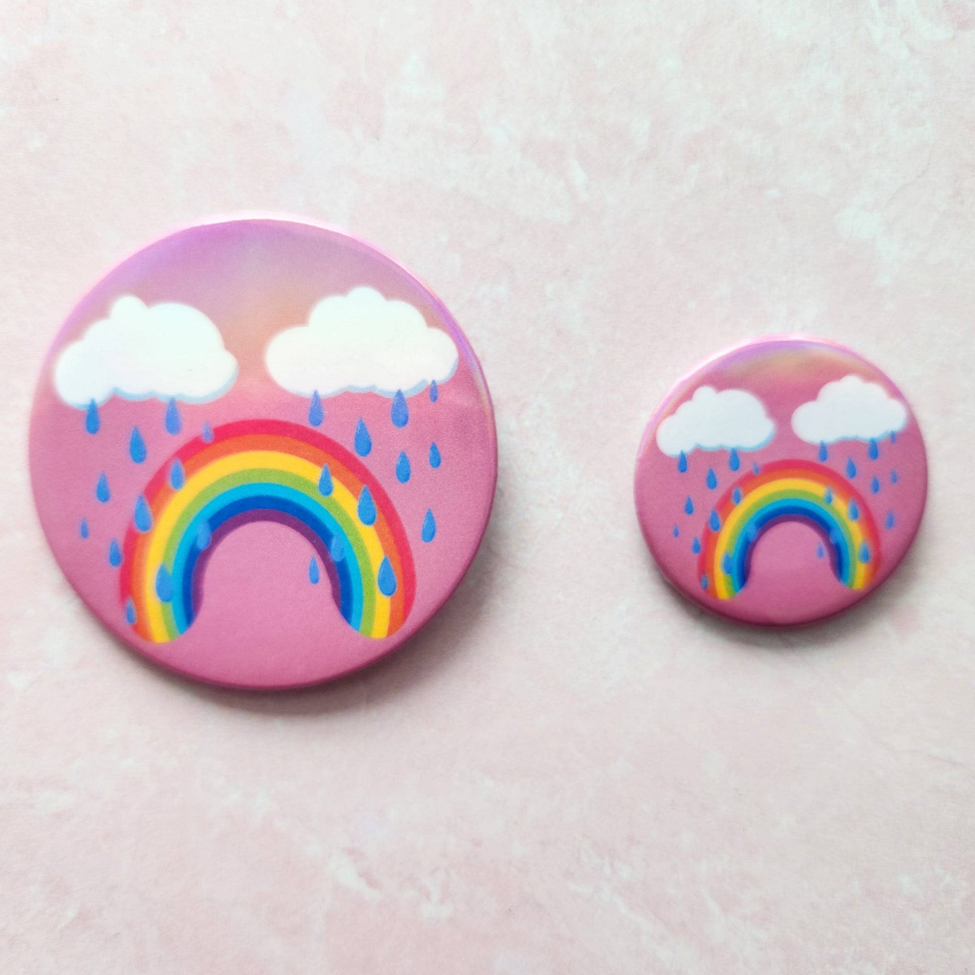 Bittersweet rainbow holographic button pin with a pink background, featuring a crying rainbow under two clouds with raindrops, one larger next to a smaller