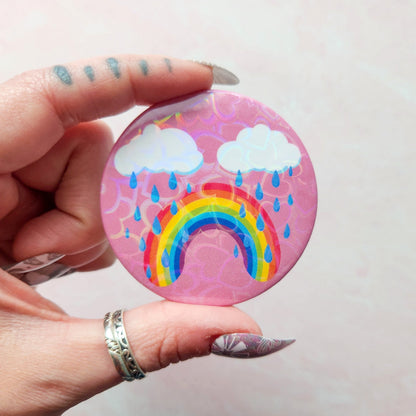 Bittersweet rainbow holographic button pin with a pink background, featuring a crying rainbow under two clouds with raindrops, held between fingers with decorative nail art.