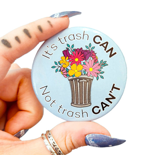 Closeup of a hand with blue nail art holding a light blue motivational button pin that reads "It's trash CAN Not trash CAN'T" with a drawing of a trash can filled with colorful flowers.