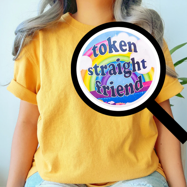 Person wearing a yellow shirt with a close-up view of a colorful 'Token Straight Friend' holographic pin featuring a bold rainbow design and playful text, perfect for pride events and allyship.