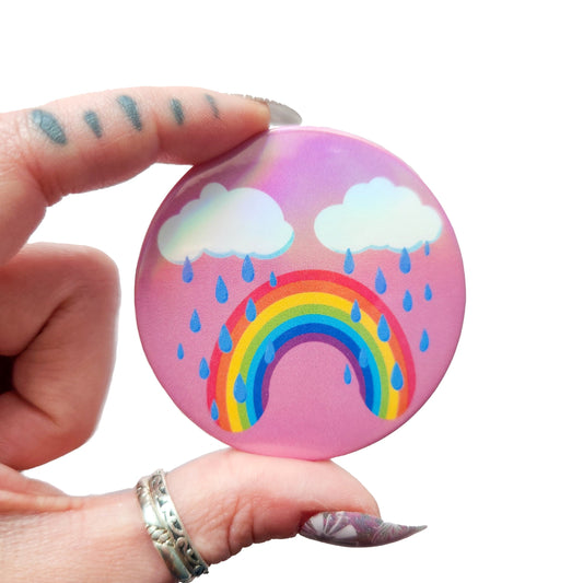 Bittersweet rainbow holographic button pin with a pink background, featuring a crying rainbow under two clouds with raindrops